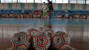 Μια ομάδα παιδιών κάθεται σε ένα κλειστό γήπεδο μπασκετ και παρακολουθεί ένα νεαρό αγόρι όπου έχει δεμένα τα μάτια του και με την καθοδήγηση του προπονητή ετοιμάζεται να ρίξει με μια μπάλα ποδοσφαίρου σε άλλες μπάλες που είναι τοποθετημένες με τη μορφή κορινών μποουλινγκ